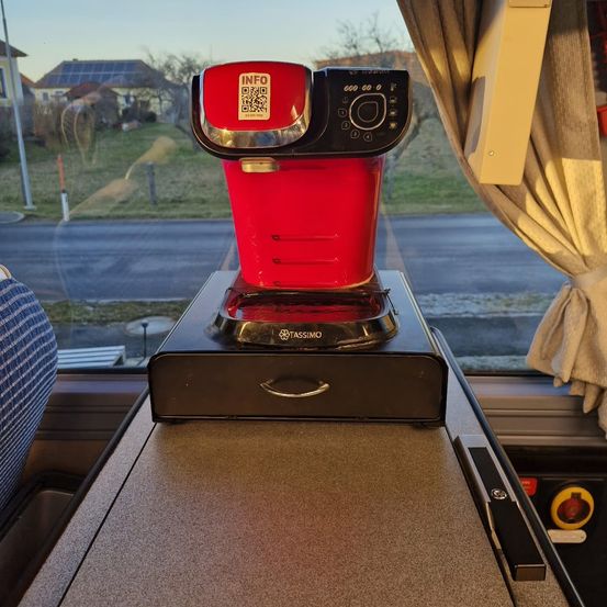 Eine rote Tassimo-Kaffeemaschine steht auf einem Tisch in einem Fahrzeug mit Blick aus dem Fenster auf eine ländliche Landschaft.