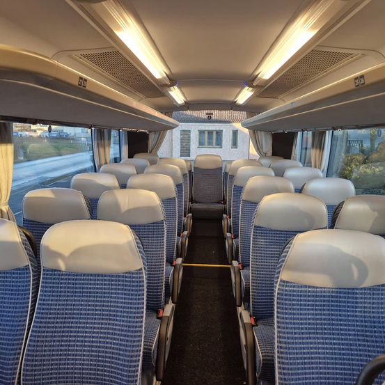Das Innere eines Busses mit blauen und weißen Sitzen, Reihen von Sitzen auf beiden Seiten und einem Gang. Die Fenster zeigen ein Gebäude und Bäume.