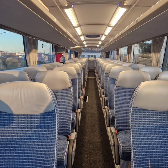 Ein leerer langer Bus mit blauen und weißen Sitzen und Lampen an der Decke. Der Bus hat Fenster auf beiden Seiten und der Außenblick zeigt ein Gebäude und Bäume.