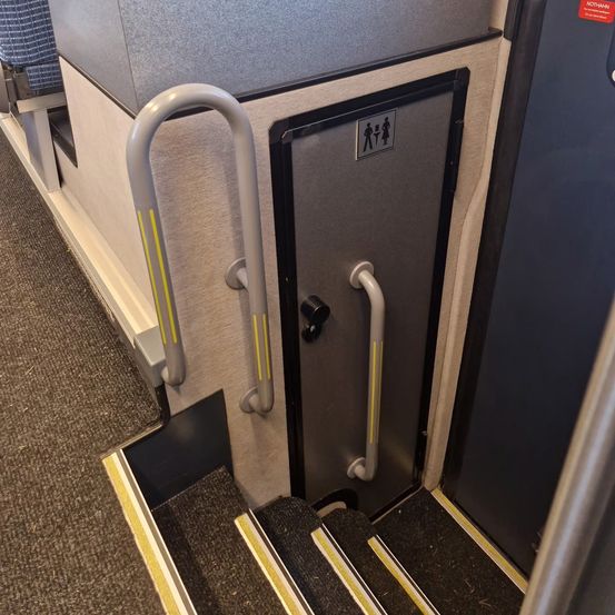 Innenansicht eines Fahrzeugs mit einer Tür, die ein WC-Schild trägt, zwei Handläufen und Stufen mit gelben Streifen an den Kanten.