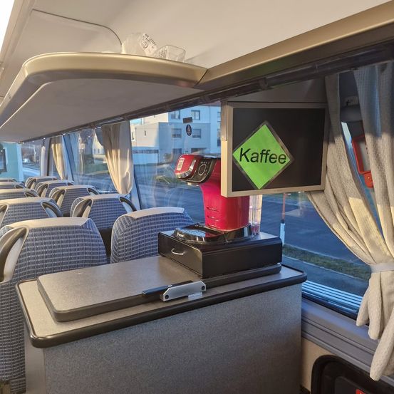 Der leere Innenraum eines Busses mit Reihen blauer Sitze. Eine Kaffeemaschine mit einem Schild, das 'Kaffee' sagt, steht auf einem Tisch in der Nähe des Fensters.