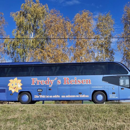 Ein blauer Doppeldecker-Bus namens 'Fredy's Reisen' steht vor einem Feld mit Gras und Bäumen. Der Bus hat ein fröhliches Sonnenlogo und deutschen Text.