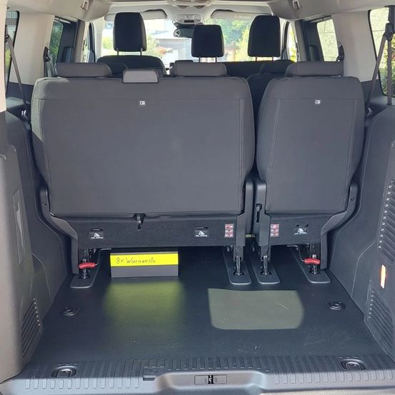 Der Innenraum eines Vans zeigt die umgeklappten Rücksitze mit einem gelben Schild, das '8x Warmweste' besagt. Die Sitze sind grau mit einem Metallgriff an der Seite. Der Boden ist schwarz mit einer silbernen Linie.