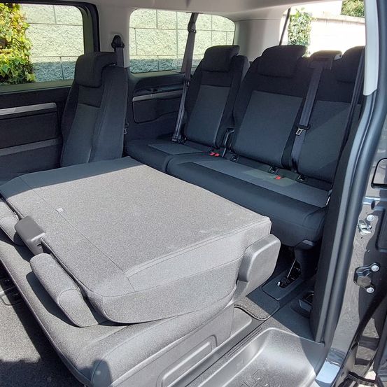 Das Innere eines Fahrzeugs zeigt vier graue Sitze mit Sicherheitsgurten, einen klappbaren Sitz vorne und eine teilweise geöffnete Tür.