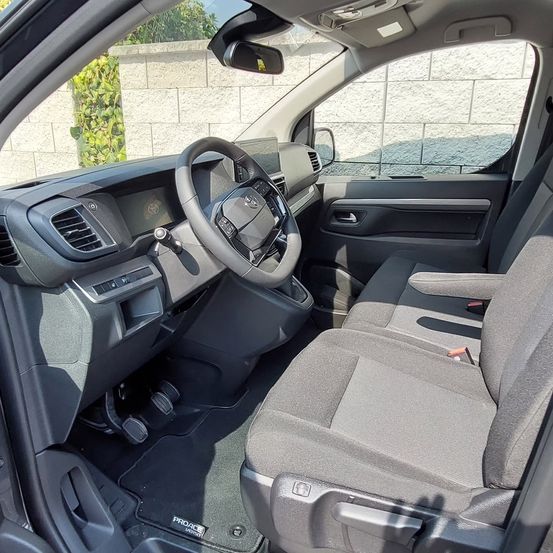 Innenraum eines Toyota Proace City Verso Vans. Der Fahrersitz und das Lenkrad sind sichtbar, ebenso ein Teil des Armaturenbretts. Die Rücksitze sind grau und haben eine matte Oberfläche.