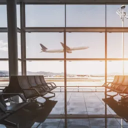 Ein Flughafenterminal mit großen Glasfenstern, die ein startendes Flugzeug zeigen. Im Inneren sind Reihen leerer Sitze zu sehen, die das Sonnenlicht auf dem gefliesten Boden reflektieren.