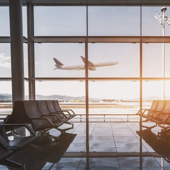 Ein Flughafenterminal mit großen Glasfenstern, die ein startendes Flugzeug zeigen. Im Inneren sind Reihen leerer Sitze zu sehen, die das Sonnenlicht auf dem gefliesten Boden reflektieren.