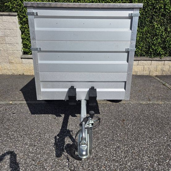 Ein silberner Cargo-Anhänger mit Metallrahmen steht auf einem Kiesweg, mit einem Schatten auf dem Boden.