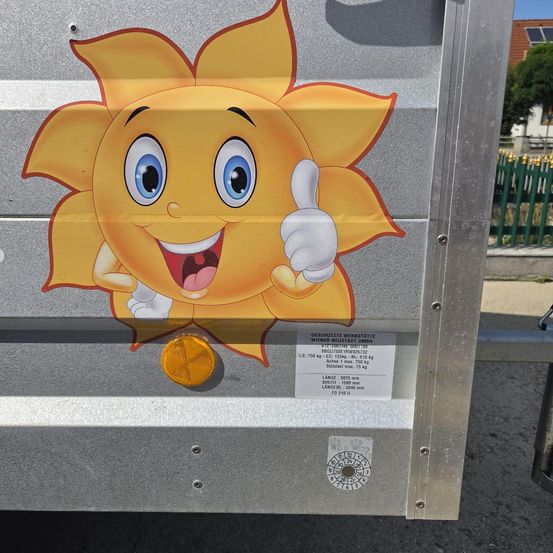 Ein Metallanhänger hat einen Sonnenaufkleber mit Daumen hoch, darunter ein Etikett mit Details wie Gewicht und Abmessungen.