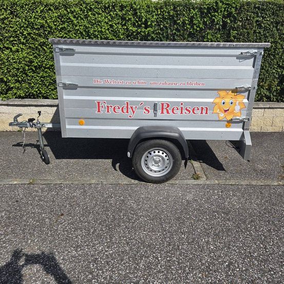 Ein silberner Anhänger mit der Aufschrift 'Fredys Reisen' an der Seite, geparkt auf einer Straße mit einem Schatten auf dem Boden.