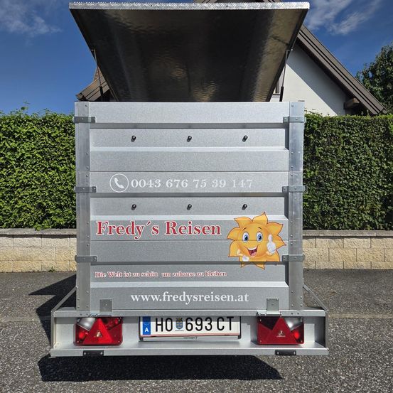 Die Rückseite eines silbernen Anhängers mit 'Fredy's Reisen' darauf, mit einer Cartoon-Sonne und Daumen hoch. Dahinter ein Haus mit braunem Dach und einer Hecke. Das Kennzeichen lautet 'HO 693 CT'.