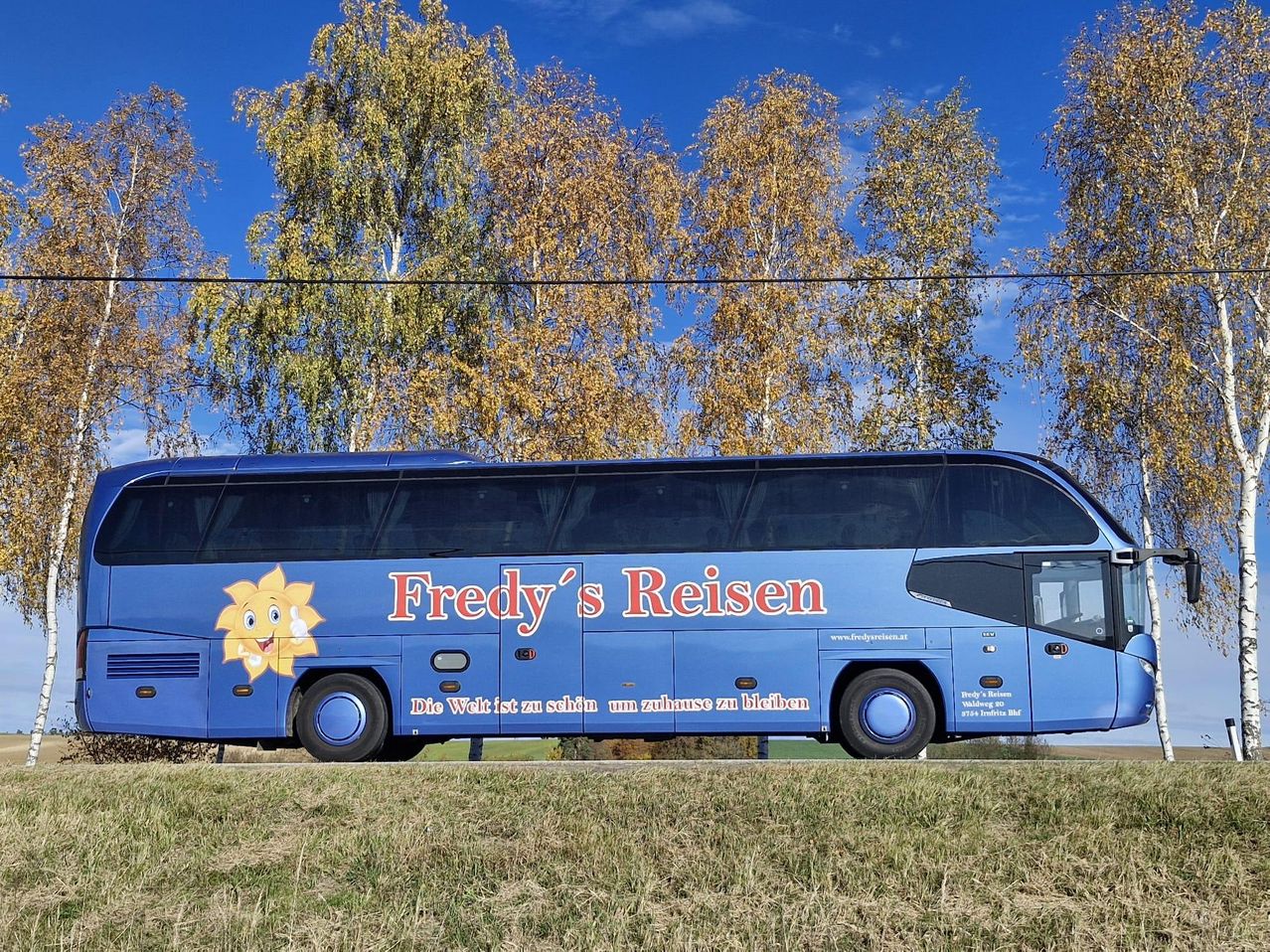Ein blauer Doppeldeckerbus mit Fredy's Reisen auf der Seite steht am Straßenrand, mit einigen Bäumen im Hintergrund.