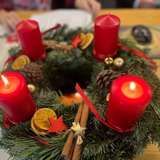 Ein Kranz mit roten Kerzen, Tannenzapfen, Zimtstangen und Orangenscheiben auf einem Holztisch. Der Kranz ist mit einem roten Band und einem Stern geschmückt.