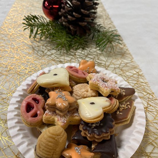 Ein Teller mit verschiedenen dekorierten Weihnachtskeksen, darunter Herz-, Stern- und Tierformen, steht auf einem Tisch mit goldenem Tuch. Im Hintergrund befinden sich eine Tannenzapfen und ein roter Weihnachtsbaumschmuck.