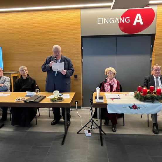 Ein Mann steht an einem Podium mit Mikrofon und Papier, während andere an Tischen sitzen. Dahinter befindet sich eine Holzwand mit dem Schild 'Eingang A' und doppelten Türen.