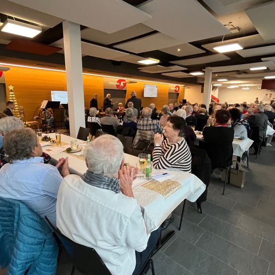 Eine Gruppe älterer Menschen sitzt an einem langen Tisch in einem Raum. Sie klatschen und schauen auf die Bühne. Links steht ein Weihnachtsbaum.