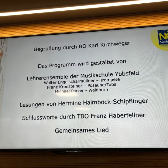 Ein Präsentationsfolie auf einem Bildschirm mit gelbem Rand. Es listet eine Begrüßung durch BO Karl Kirchweger, Programmdetails und Namen von Mitgliedern des Musikschulensembles auf.