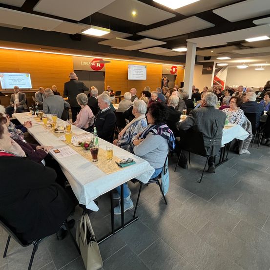 Ein Raum mit mehreren Tischen und Stühlen. Menschen sitzen um Tische herum, einige stehen und eine Person präsentiert an einem Podium. Der Raum hat eine Projektionsleinwand und Lampen an der Decke.