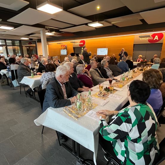 Ein Raum mit langen Tischen, an denen mehrere ältere Menschen sitzen. Auf den Tischen stehen Gläser und Teller. Im Hintergrund befindet sich ein Podium mit einem Bildschirm.