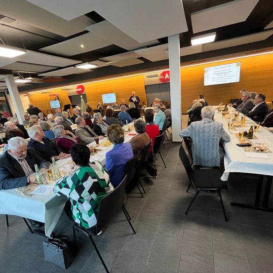 Eine Gruppe älterer Menschen sitzt um einen langen Tisch herum und nimmt an einer Versammlung teil. Der Raum hat ein Eingangsschild und eine Projektionsleinwand. Viele Tische haben Teller, Gläser und Essen darauf.