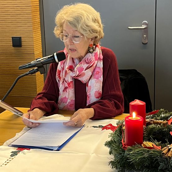 Eine ältere Frau mit Brille und einem Blumenschal sitzt an einem Tisch mit einem Mikrofon und liest ein Papier. Zwei rote Kerzen und ein Kranz sind auf dem Tisch. Dahinter befindet sich eine Holzwand und eine graue Tür.