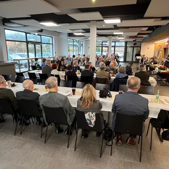 Eine große Gruppe von Menschen sitzt um Tische in einem Speisesaal. Einige Tische haben Essen und Getränke darauf. Es sind Autos vor den Fenstern geparkt.