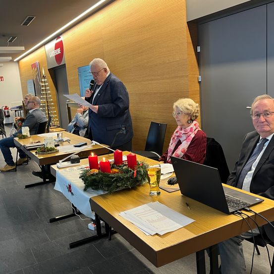 Ein Mann steht an einem Tisch mit Mikrofon und Papieren und spricht zu einer Gruppe von Menschen, die um einen Tisch mit einer Girlande, Kerzen und einem Laptop sitzen.