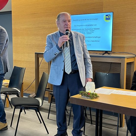 Ein älterer Mann in einem Anzug steht vor einem Schreibtisch, hält ein Mikrofon in der Hand und hält vermutlich eine Rede. Hinter ihm ist ein Bildschirm mit Text. Ein hölzerner Schreibtisch mit einer Kerze und Papieren steht vor ihm, mit Stühlen und einem Tisch an der Seite.