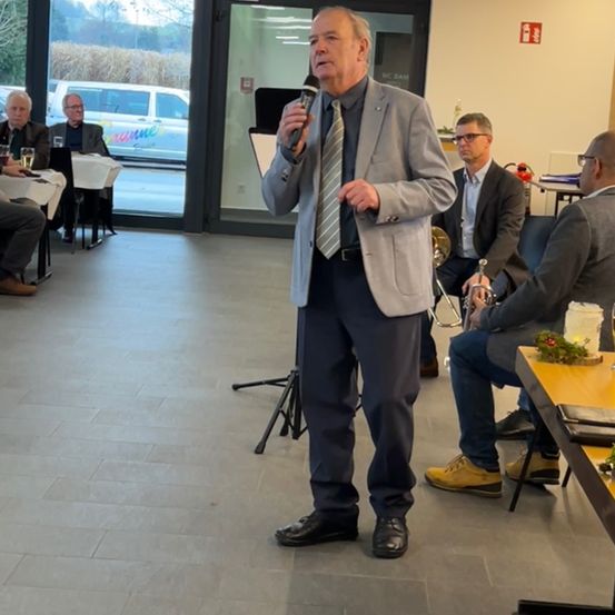 Ein Mann steht und spricht in ein Mikrofon in einem Raum. Hinter ihm sitzen drei Personen, eine mit einer Trompete. Tische, Stühle und eine Glastür sind im Raum.