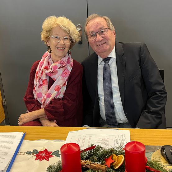 Eine ältere Frau mit Brille und einem Blumenschal sitzt neben einem Mann in Anzug und Krawatte, beide lächeln für die Kamera. Sie sitzen an einem Tisch mit Papieren, Kerzen und einer Maus.