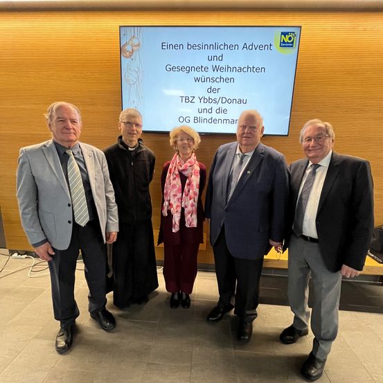 Fünf formell gekleidete Personen stehen vor einer Projektionsleinwand mit einer festlichen Botschaft, möglicherweise bei einer Feierlichkeit.