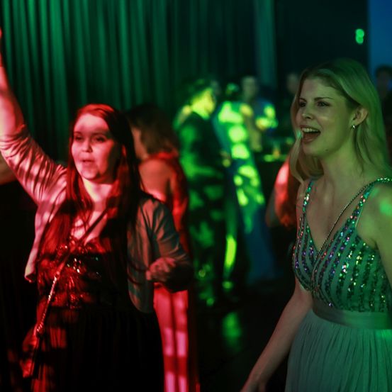 Zwei Frauen tanzen bei einer lebhaften Veranstaltung. Eine in einem grünen Kleid lächelt und hebt ihren Arm. Die andere in einem roten Kleid steht neben ihr. Sie sind von einer Menschenmenge umgeben, und der Raum ist mit grünen Lichtern beleuchtet.