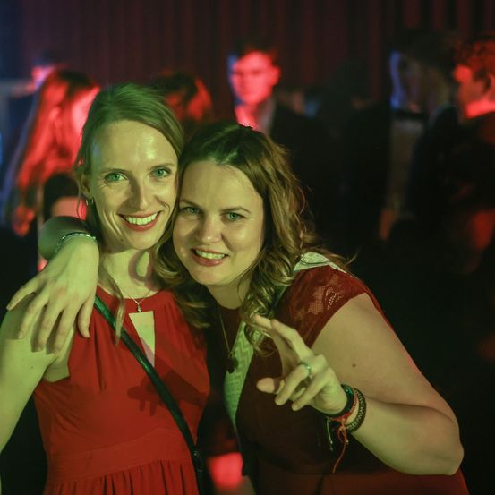 Zwei Frauen in roten Kleidern posieren für ein Foto auf einer Party mit roter Beleuchtung und anderen Gästen im Hintergrund.