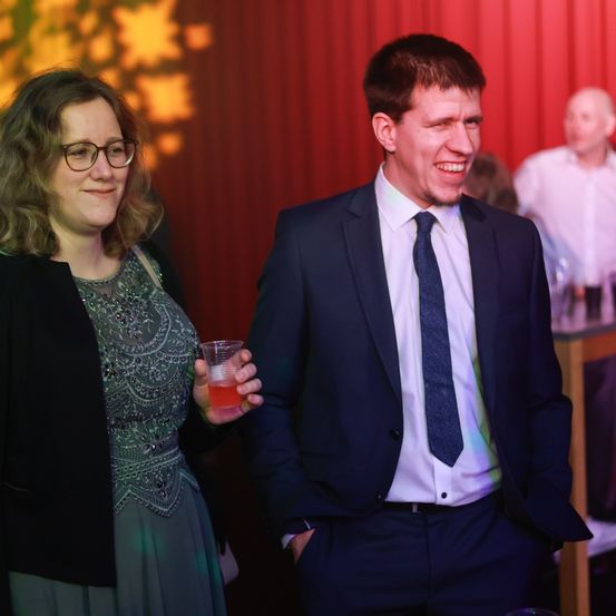 Eine Frau und ein Mann stehen lächelnd zusammen in einem formellen Umfeld. Die Frau hält ein Glas mit roter Flüssigkeit. Hinter ihnen ist ein weiterer Mann in einem weißen Hemd teilweise sichtbar.