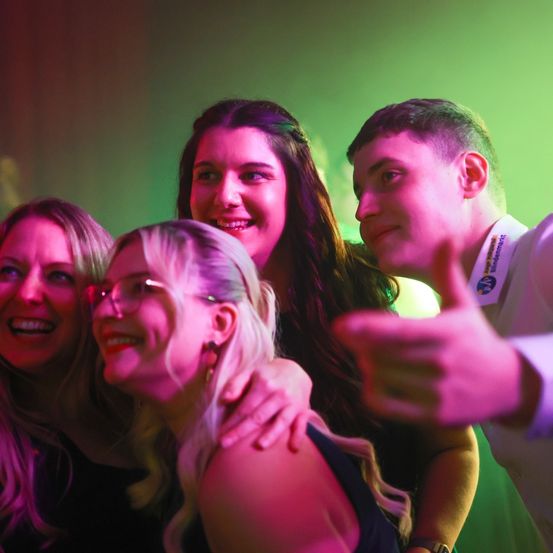 Vier Personen lächeln in die Kamera. Sie scheinen auf einer Party zu sein, unter rosa und grünem Licht. Eine Frau hat ihren Arm um eine andere Frau gelegt.