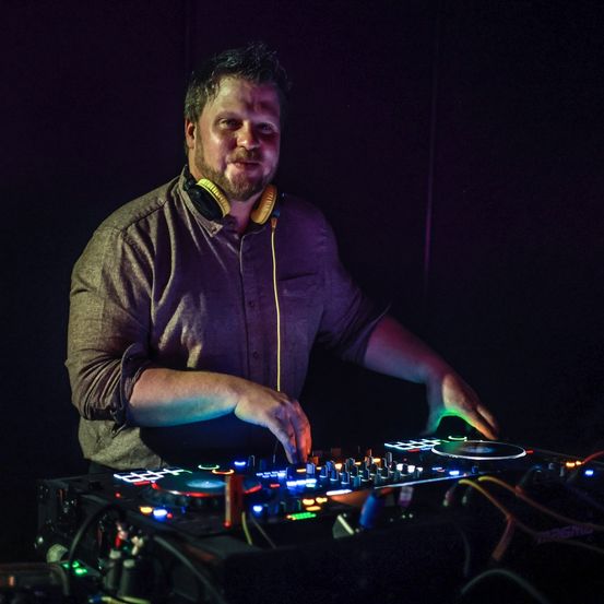 Ein Mann steht hinter einem DJ-Setup mit bunten Lichtern, trägt Kopfhörer und ein lila Hemd und wird von einem Scheinwerfer beleuchtet.