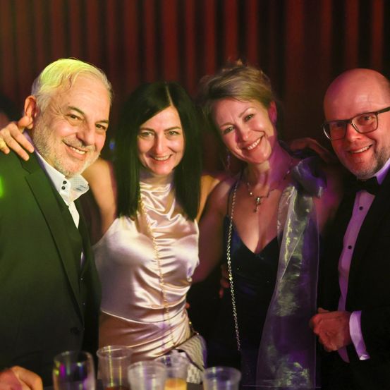 Vier Menschen auf einer Party, lächelnd. Zwei Frauen und zwei Männer. Die Frau links trägt ein rosa Kleid und die Frau rechts trägt einen lila Schal. Die Männer tragen Anzüge.