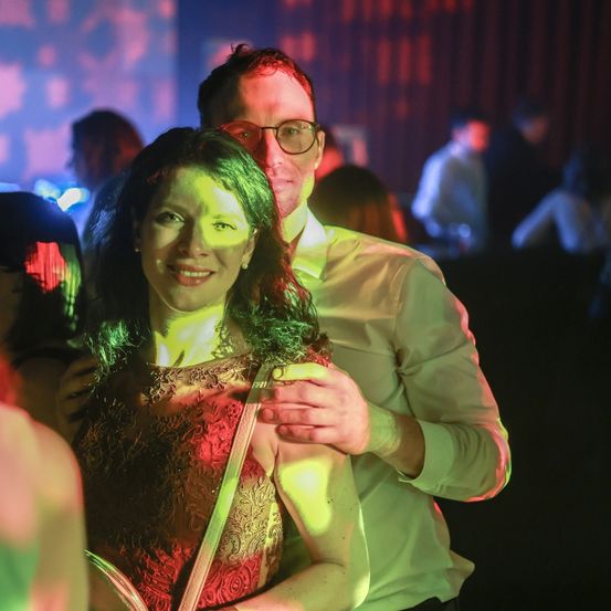 Ein Paar in einem schwach beleuchteten Raum, lächelt und posiert für ein Foto. Die Frau trägt ein rotes Kleid, und der Mann, eine Brille. Unscharfe Menschen und bunte Lichter im Hintergrund.