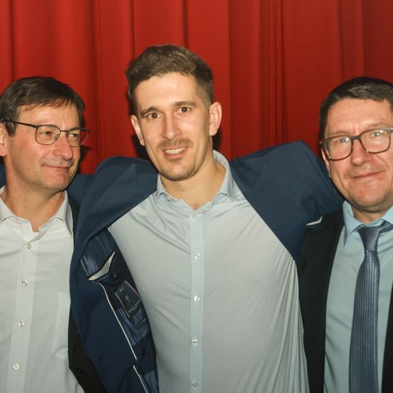 Drei Männer in Anzughose posieren vor einem roten Vorhang. Der Mann in der Mitte lächelt, während er die Schultern der anderen umarmt.