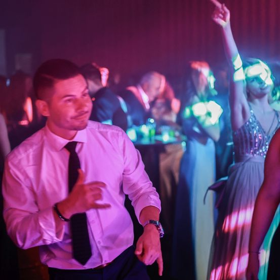 Ein Mann in einem weißen Hemd und schwarzer Krawatte tanzt in einem Club, mit einer Frau, die in der Nähe ihre Hände hebt. Die Atmosphäre ist von rosa Lichtern erleuchtet.