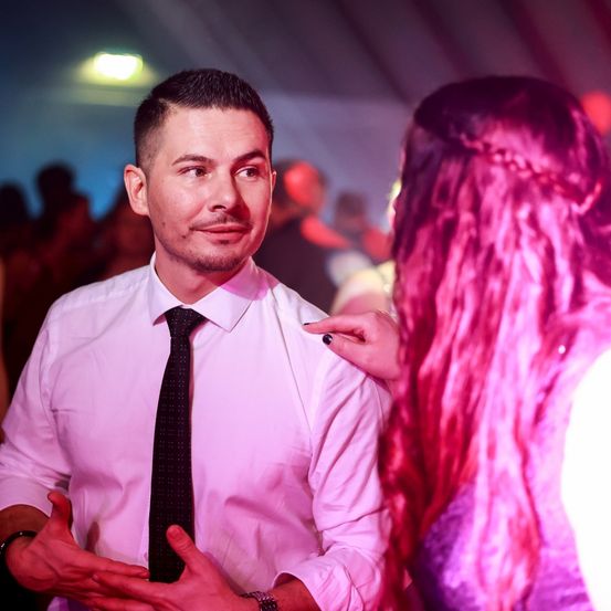 Ein Mann in einem weißen Hemd und schwarzer Krawatte spricht mit einer Frau mit rosa Haaren in einer gesellschaftlichen Umgebung.