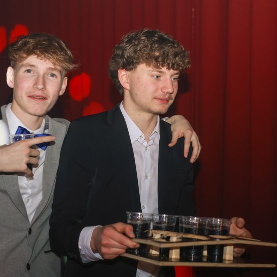 Zwei junge Männer in Anzugkleidung, einer mit einem Tablett mit Getränken, posieren für ein Foto in einem rot beleuchteten Raum.
