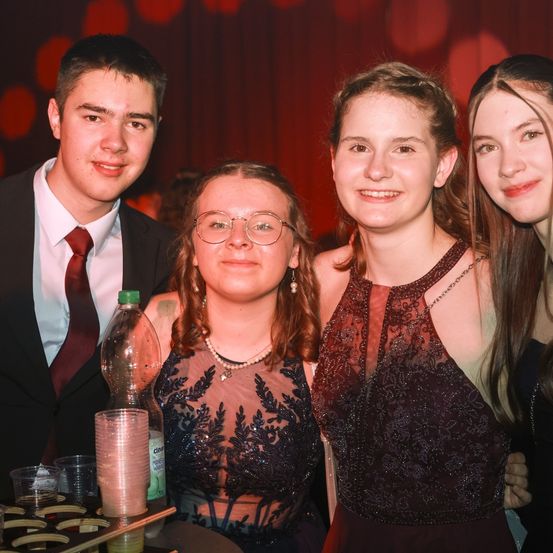 Vier Personen posieren für ein Foto bei einer Veranstaltung. Ein junger Mann in Anzug und Krawatte und drei Frauen, zwei in Kleidern und eine mit Brille. Ein Tisch mit Bechern und einer Flasche steht vor ihnen.