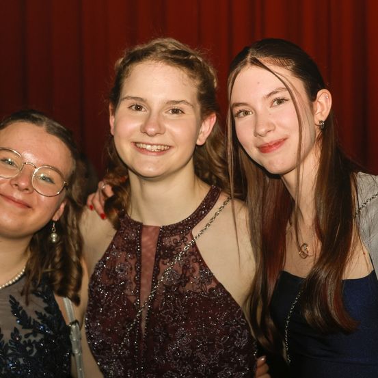 Drei Frauen posieren vor einem roten Vorhang für ein Foto. Die Frau in der Mitte trägt ein burgunderrotes Kleid und eine Halskette. Die anderen beiden Frauen lächeln.