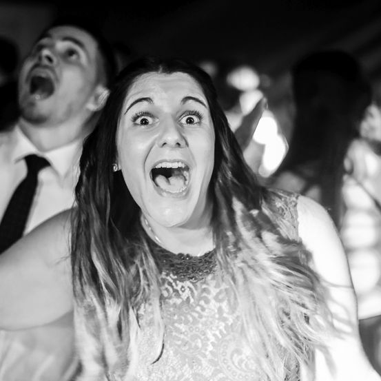 Eine Frau mit langen Haaren und einem Kleid schreit auf einer Party. Hinter ihr steht ein Mann mit Krawatte und eine Frau mit Pferdeschwanz.