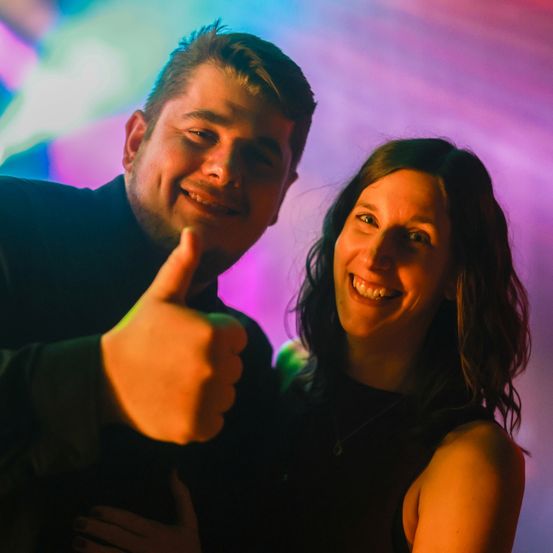 Ein Mann und eine Frau lächeln und posieren für ein Foto vor einem farbenfrohen Hintergrund. Der Mann gibt einen Daumen nach oben.