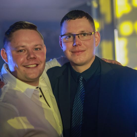 Zwei Männer lächeln und posieren für ein Foto. Der Mann auf der linken Seite trägt ein helles Hemd. Der Mann auf der rechten Seite trägt einen dunklen Anzug und eine Krawatte. Der Hintergrund ist schwach beleuchtet.