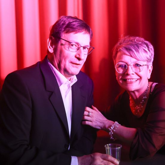 Ein älteres Paar, beide mit Brille, posiert für ein Foto. Der Mann trägt einen schwarzen Anzug, und die Frau trägt eine dunkle Bluse und eine Halskette. Sie sitzen an einem Tisch und lächeln.
