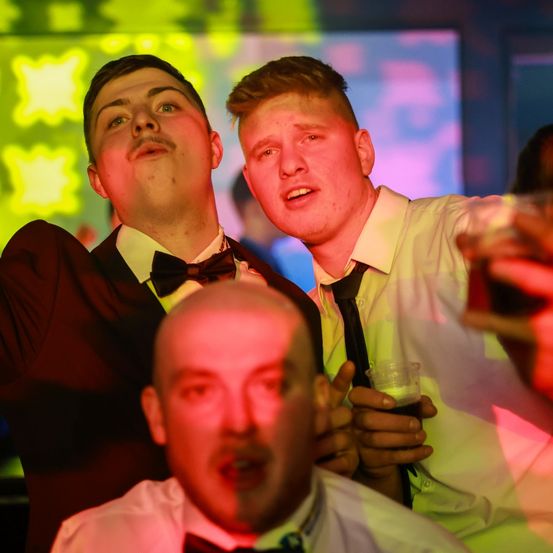 Drei Männer auf einer Party, einer hält ein Getränk, ein anderer macht lustige Gesichter, mit einem farbenfrohen Hintergrund und gelben Sternen.