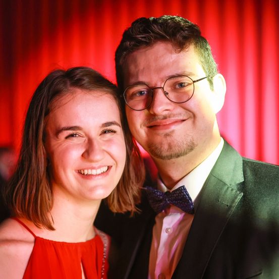 Ein Mann und eine Frau lächeln und posieren für ein Foto. Sie stehen eng beieinander. Der Mann trägt einen Anzug und eine Brille. Die Frau trägt ein rotes Kleid. Der Hintergrund ist rot.